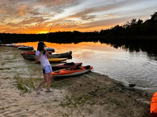 A person casts a fishing line into a calm lake at sunset, surrounded by colorful kayaks on the shore.