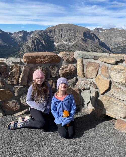 Two girls in winter hats kneel by a stone wall, smiling with mountains in the background on a sunny day.