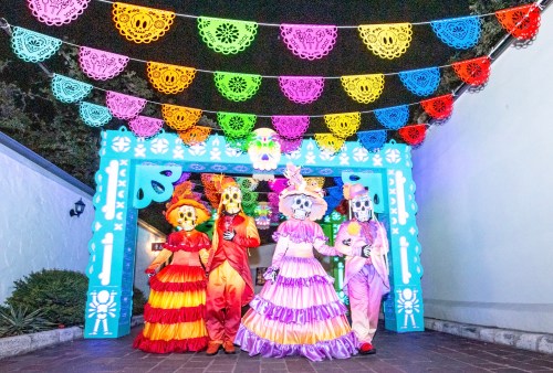 Four people in colorful costumes and skull masks stand under vibrant papel picado decorations at a festive event.