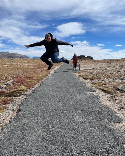 A person jumps joyfully on a path in a scenic landscape, with another figure walking in the background.