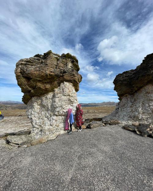 Two women in colorful attire stand between two large rock formations under a blue sky with scattered clouds.