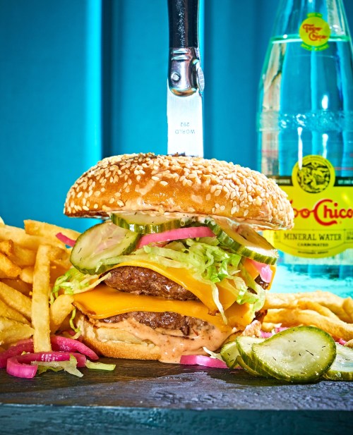 A juicy cheeseburger with lettuce, pickles, and onions, served with fries and a bottle of mineral water.