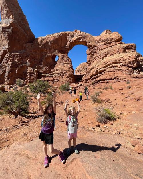 Two children with raised hands pose in front of a large rock arch in a desert landscape, with other hikers in the background.
