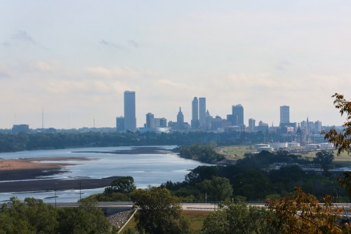 A panoramic view of a city skyline with a river in the foreground and greenery along the banks.