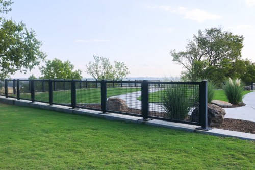 A black metal fence surrounds a grassy area with trees and rocks, leading to a winding path under a blue sky.