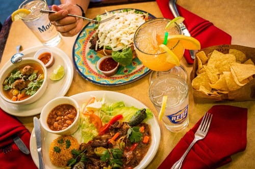 A vibrant table spread featuring tacos, beans, salad, chips, and drinks, with a hand reaching for a plate.