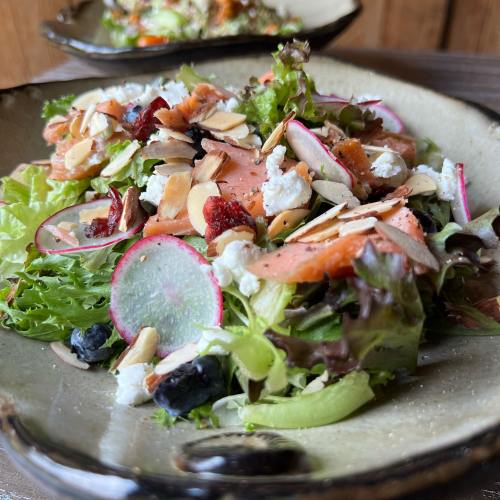 A fresh salad with greens, smoked salmon, radishes, almonds, and goat cheese, served in a rustic bowl.