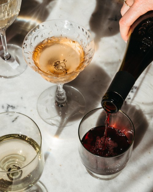 A hand pours red wine into a glass, surrounded by champagne and white wine glasses on a marble surface.