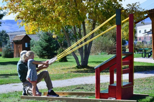 A child and an adult play with a large rubber band slingshot in a park surrounded by trees and colorful autumn foliage.