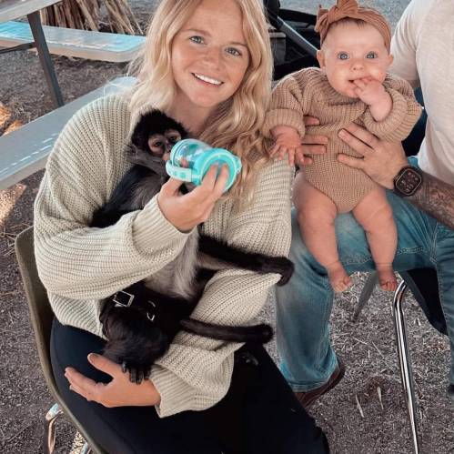 A woman holds a baby monkey while sitting next to a child, both smiling in a casual outdoor setting.