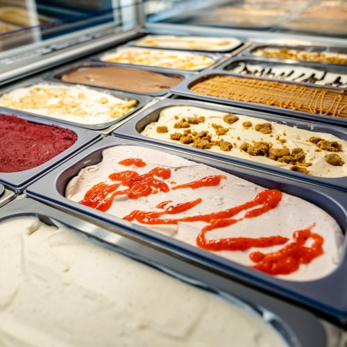 A variety of gelato flavors in metal containers, including red fruit, chocolate, and creamy vanilla with toppings.