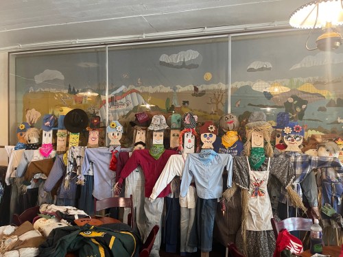 A row of colorful scarecrows dressed in various outfits, displayed against a scenic mural backdrop.