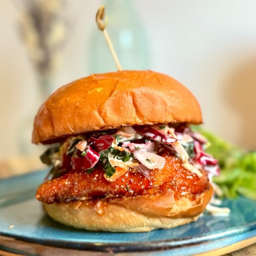 A crispy fried chicken sandwich topped with coleslaw, served on a toasted bun with a toothpick.