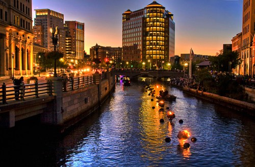 A cityscape at dusk featuring a river with floating lights, buildings, and a lively waterfront scene.