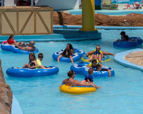 A group of people relaxes on inner tubes in a blue water park pool on a sunny day.