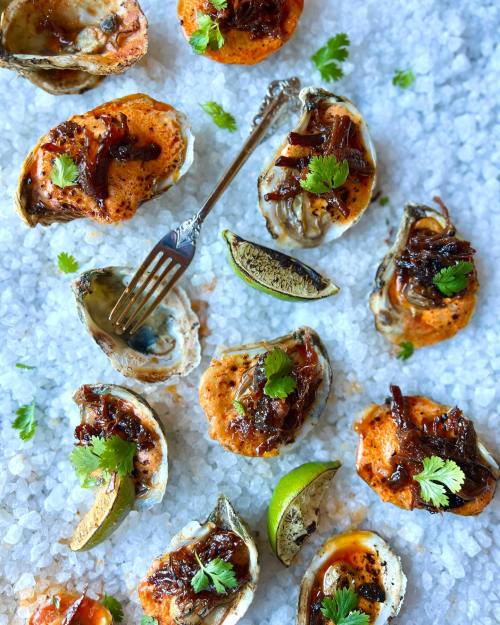 A platter of baked oysters topped with caramelized onions, garnished with cilantro and lime wedges on a bed of ice.
