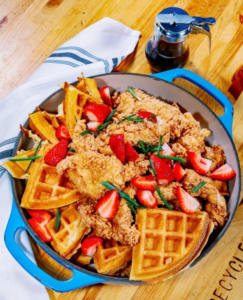 A bowl filled with fried chicken, waffles, strawberries, and green onions, with syrup in a jar beside it.