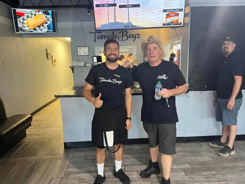 Two men stand in a restaurant, smiling and posing for a photo. A menu is visible in the background.