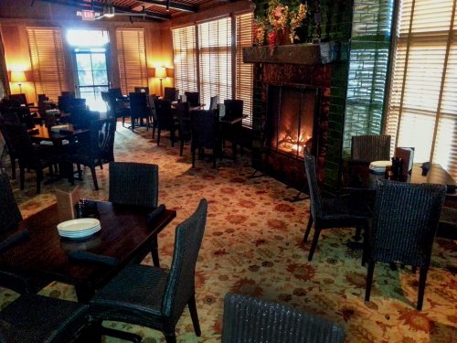 Cozy restaurant interior with tables, chairs, and a fireplace, featuring large windows and warm lighting.