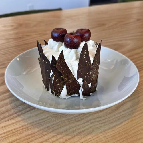 A round cake with chocolate shards, whipped cream, and three cherries on top, served on a white plate.