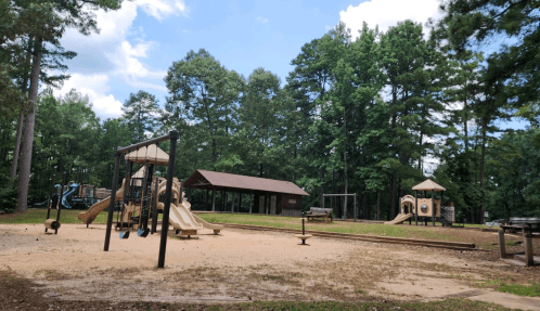 A playground with slides and climbing structures, surrounded by trees and a picnic shelter in a sunny park.
