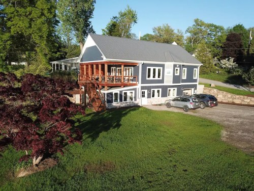 A modern two-story house with a wooden deck, surrounded by greenery and parked cars in a gravel driveway.