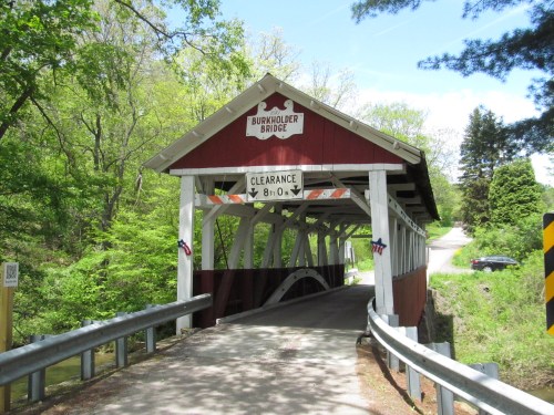 A red and white covered bridge with a sign indicating clearance, surrounded by lush green trees and a winding road.