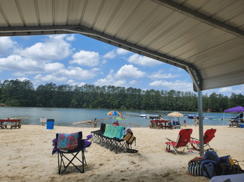 A sandy beach scene with chairs, umbrellas, and people enjoying a lake under a blue sky with fluffy clouds.