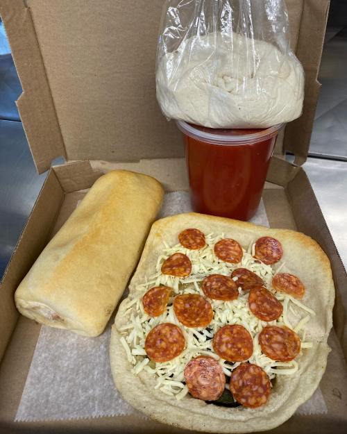 A takeout box containing a pizza with pepperoni, a bread roll, and a cup of red drink, all neatly arranged.