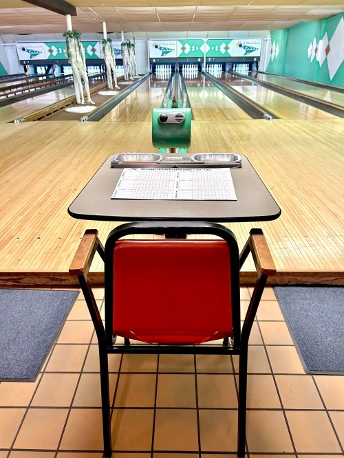 A bowling alley view featuring a table with a score sheet and a red chair, overlooking the lanes.
