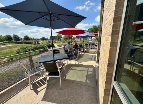 Outdoor seating area with umbrellas, tables, and chairs, overlooking a green space under a blue sky with clouds.