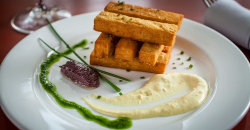 A plate featuring stacked golden tofu, garnished with green sauce and a dark spread, alongside a creamy dip.