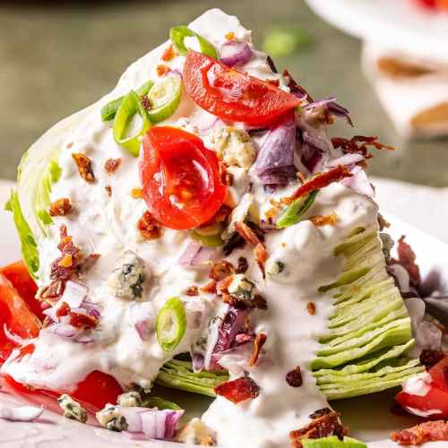 A wedge salad topped with tomatoes, bacon, green onions, and blue cheese dressing on a white plate.
