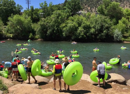 People enjoying a sunny day floating on green inner tubes in a river surrounded by trees and rocks.