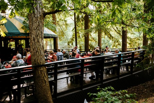 A group of people dining outdoors on a wooden deck surrounded by trees, enjoying a meal in a natural setting.
