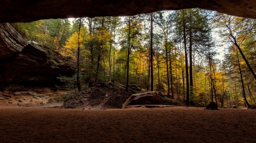 A scenic view from inside a cave, showcasing vibrant autumn trees and a sandy floor illuminated by soft sunlight.