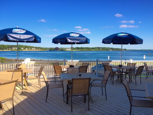 Outdoor seating area with tables and umbrellas overlooking a calm waterfront and blue sky.