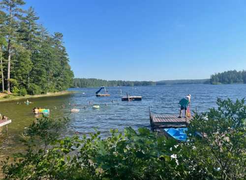 A serene lake scene with people swimming, a dock, and kayaks surrounded by lush green trees under a clear blue sky.