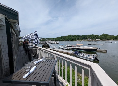 View of a marina with boats, featuring a restaurant deck with striped umbrellas and a scenic waterfront.