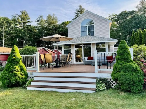 A charming house with a deck, patio furniture, and lush greenery, surrounded by trees and a clear blue sky.