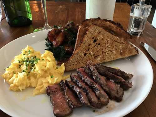 A plate featuring scrambled eggs, sliced steak, toasted bread, and sautéed greens, set on a wooden table.