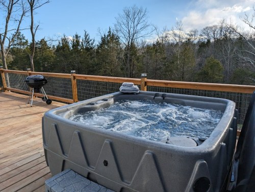 A hot tub on a wooden deck with a grill nearby, surrounded by trees and a clear blue sky.