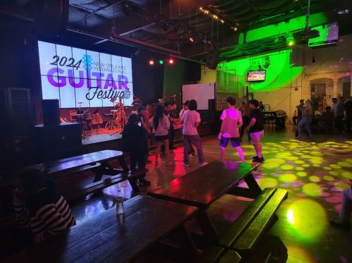 A lively indoor venue with people dancing, a stage for the 2024 Guitar Festival, and colorful lights illuminating the space.