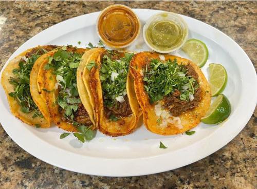 A plate of four tacos topped with cilantro and onions, served with lime wedges and two sauces on the side.