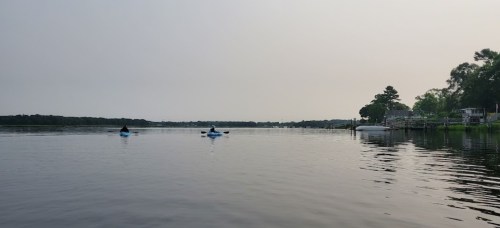 Two kayakers paddle on a calm river, with trees and a house visible along the shore under a hazy sky.