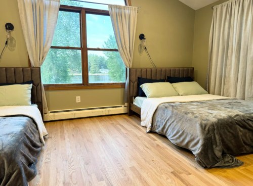 A cozy bedroom with two beds, large windows, and soft curtains, overlooking a serene outdoor view.