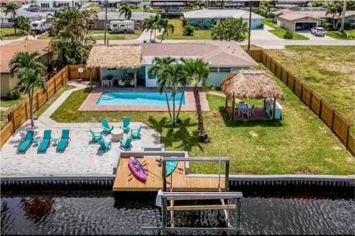 Aerial view of a backyard with a pool, lounge chairs, and a dock by a canal, surrounded by lush greenery.