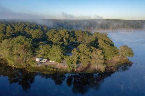 Aerial view of a serene lakeside campsite surrounded by trees and misty water at dawn.