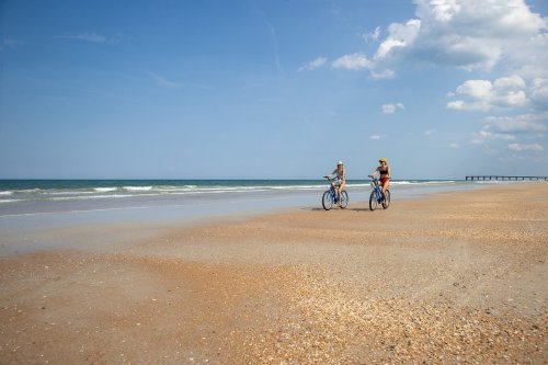 Two people ride bicycles along a sandy beach with gentle waves and a clear blue sky in the background.