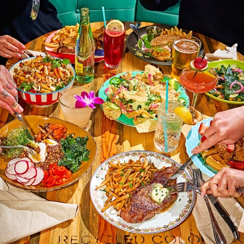 A vibrant table spread with various dishes, including salads, nachos, steak, and drinks, surrounded by hands reaching for food.